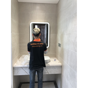 Người đứng trước gương chữ nhật có viền sáng ở phía trên chậu rửa.