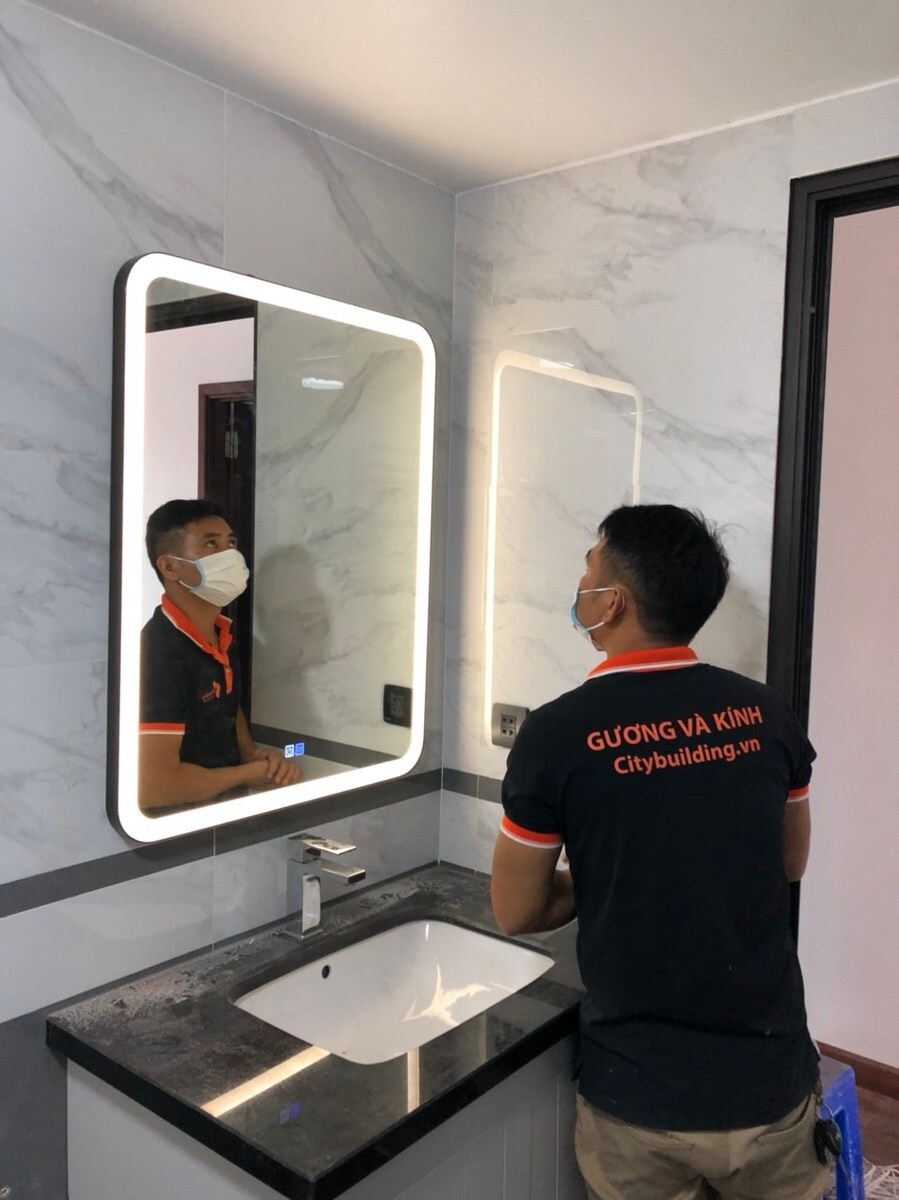 Người mặc áo Citybuilding đứng cạnh gương chữ nhật sáng viền tại lavabo