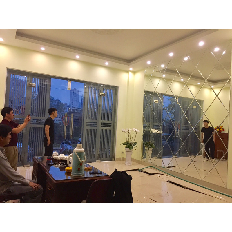 Mảng gương ghép trên tường trong phòng có bàn ghế, người và vật tư ở phía dưới.
