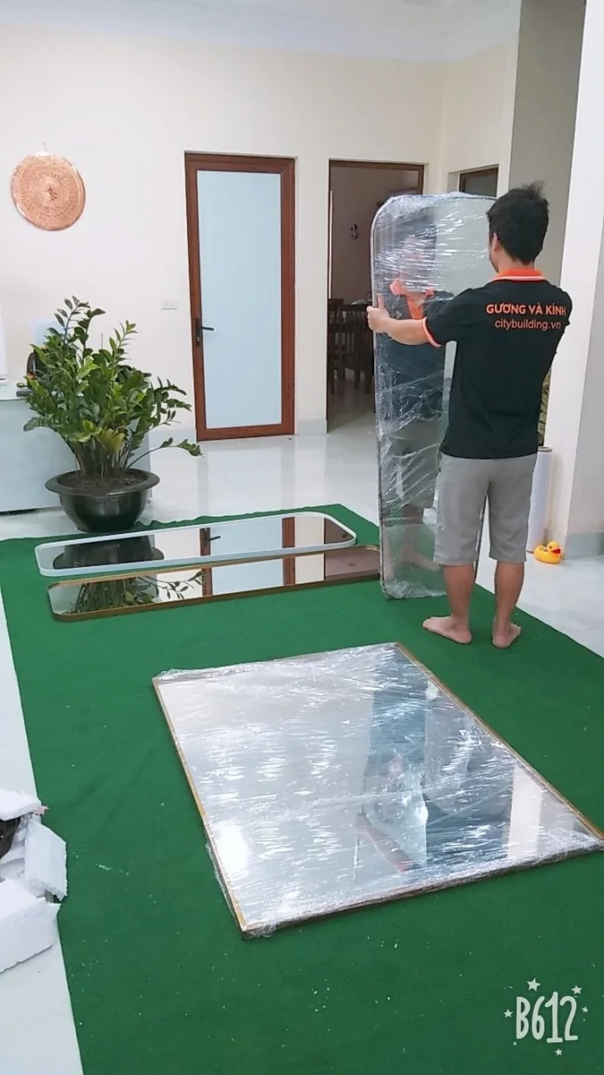 Người mặc áo có chữ citybuilding.vn giữ gương cao đã bọc màng, cạnh các gương đặt trên nền xanh
