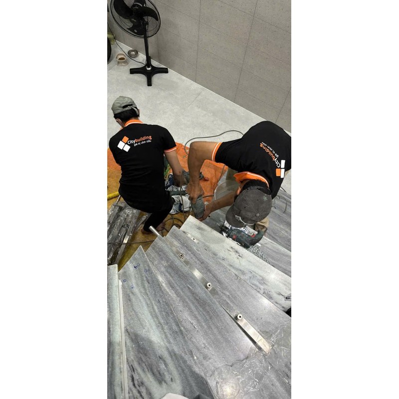 Người mặc áo Citybuilding thao tác bằng dụng cụ cầm tay tại chân bậc cầu thang, cạnh thanh liên kết đặt trên bậc.