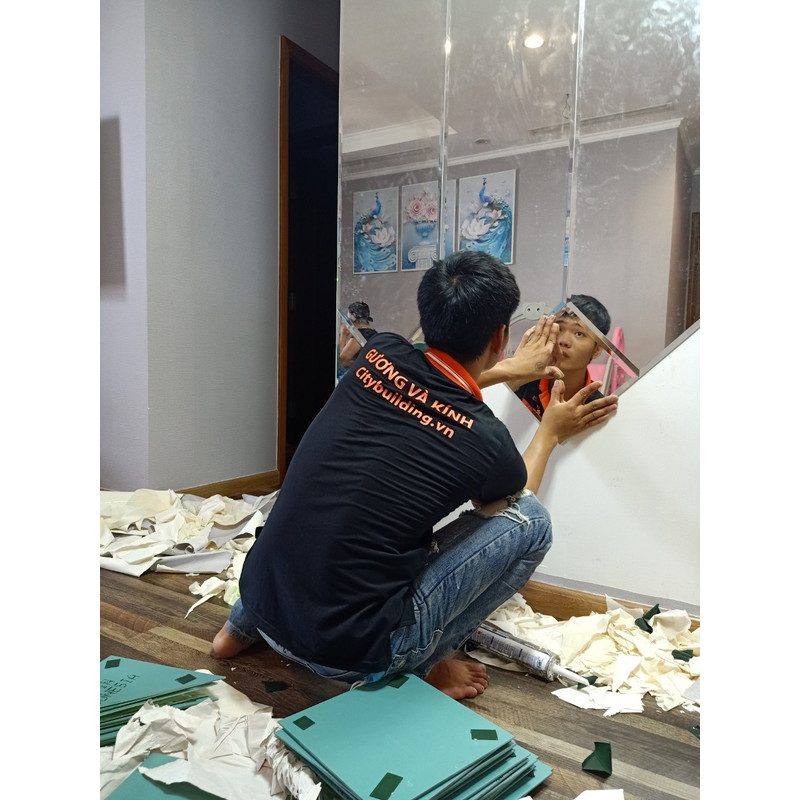 Người ngồi sát sàn, đưa tay tới một tấm gương ở phần dưới của mảng gương ghép.