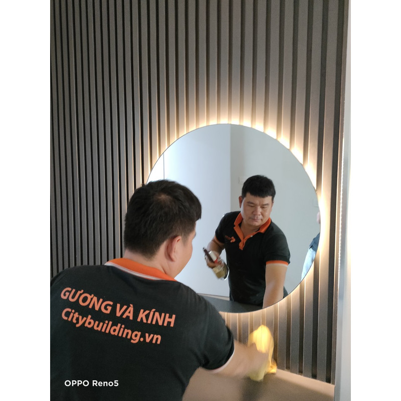 Người cầm khăn trước gương tròn có viền sáng trên vách dạng nan dọc.