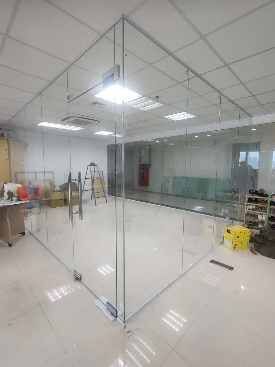 Vách kính tạo góc trong phòng có trần thả, cửa kính tay nắm dài và vật tư phía sau